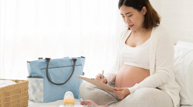 Pregnant person sits on bed with their hospital bag check list, the bag, and a few items they might choose to bring.