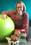 Photo: Dark teal background. Greyish plush carpeting. Smiling woman with short blond hair and swoosh bangs leans towards the camera, arm over a bright green birthing ball. She wears a colorful pink, purple and grey knit sweater that falls slightly over her hands. There is a large stack of colorful pregnancy, childbirth, breastfeeding and parenting books and a pelvis model with a Black baby doll sitting inside of it in front of her.