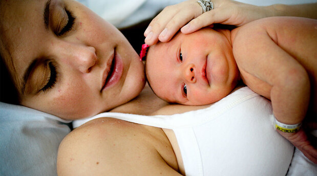 Photo of a white parent, presumably the mother, wearing a white thin strapped tank top and a large diamond wedding ring laying back on a white pillow looking down at their white naked newborn baby who is lying on the parent's chest with no clothes on with their head turned facing the camera. The baby has hospital wrist bands on the visable wrist that are yellow, with black letters and white with black letters.