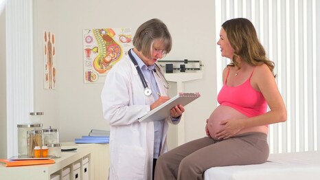 Picture: Pregnant person sittin on table in an exam room holding their belly. Doctor is is standing in front of the pregnant person looking at their clip board.