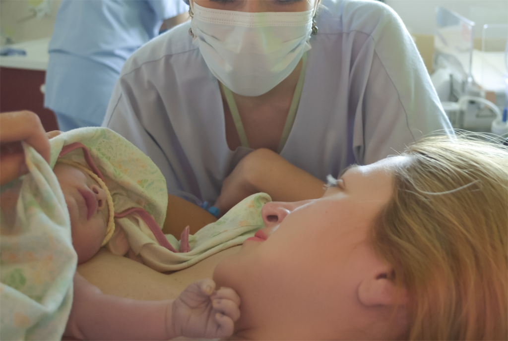 Close up of mother with baby on her chest and a doula next to her. The mother is looking at the support person smiling. The baby is covered except from it's face. The doula is wearing a surgical mask.