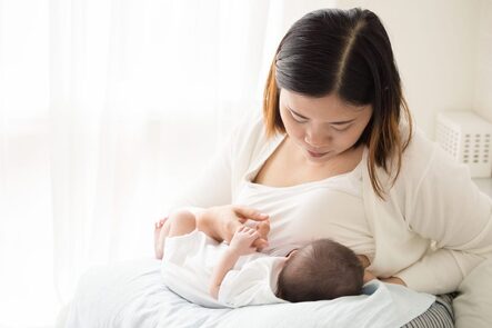 Picture of an asian mom wearing all white and her baby wearing all white on a white blanket with a white background. The mother is attempting to breastfeed her newboarn while the newboarn lays on a soft pillow on the mom's lap