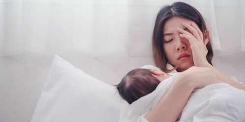 Picture of a mom with her newborn. The mom is laying on a be with her newborn in one arm. The newborn is faceing away from the camera so all you can see is the baby's hair. The postpartum mom is dealing with postpartum mood disorders and appears to be frustrated.